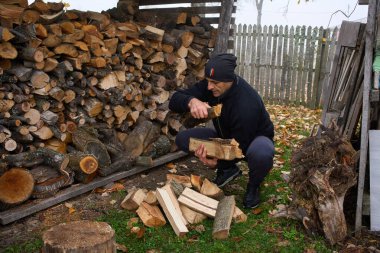 Orta yaşlı bir adam yakacak odunları baltayla kırsal kesimlerde istiflenmiş odunların yanında kırıyor.