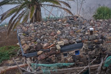 Anaga, Tenerife 'deki Hacienda Las Palmas' ta alışılmadık bir taş bina, tamamen gevşek volkanik kaya ve kayalarla kaplı, cam bloklu pencereler duvarlara kurulmuş ve arkasında Atlantik kıyısının üzerinde yükselen bir palmiye ağacı bulunan düz bir çatı.