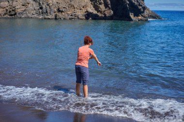 Playa Bermejo, Tenerife 'nin kara volkanik kumları üzerinde çıplak ayakla Atlantik' te yürüyen kıvırcık kızıl saçlı bir kadın engebeli kıyı kayalarına çarpıyor..