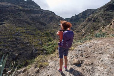 Kıvırcık kızıl saçlı ve sırt çantalı bir kadın fotoğrafçı Tenerife, Anaga 'daki dramatik Barranco de Chamorga vadisine doğru kayalık bir çıkıntıda duruyor.