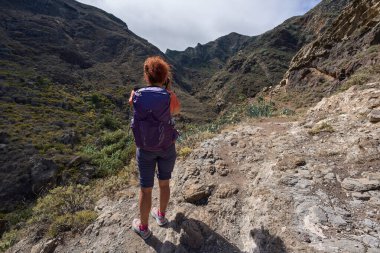 Kıvırcık kızıl saçlı ve sırt çantalı bir kadın fotoğrafçı Tenerife, Anaga 'daki dramatik Barranco de Chamorga vadisine doğru kayalık bir çıkıntıda duruyor.