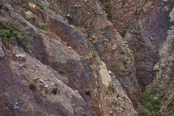 Anaga, Tenerife 'deki renkli volkanik dağ geçidi duvarları, kırmızı, turuncu, mor ve koyu demir zengini kaya çökeltilerini, aşınmış yüzeylere yapışmış, seyrek endemik çalılık bitkilerine sahip canlı, çok renkli katmanlar sergiliyor.