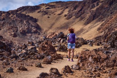 Kıvırcık kızıl saçlı ve mor sırt çantalı bir kadın volkanik lav kayalarıyla çevrili kumlu patika boyunca yürür, Tenerife, Teide platosundaki dik turuncu ve kahverengi volkanik yamaca doğru yürür.