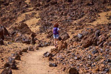Kıvırcık kızıl saçlı ve mor sırt çantalı bir kadın volkanik lav kayalarıyla çevrili kumlu patika boyunca yürür, Tenerife, Teide platosundaki dik turuncu ve kahverengi volkanik yamaca doğru yürür.