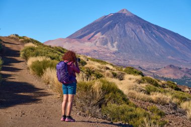 Kıvırcık kızıl saçlı ve mor sırt çantalı bir kadın, Teide Dağı 'nın yüksek konisini, açık mavi gökyüzünün altındaki Teide beyaz süpürgesiyle fotoğraflıyor.