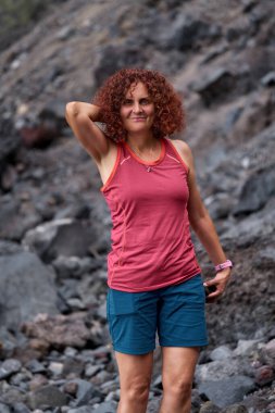 Kıvırcık kızıl saçlı, gülümseyen bir kadın. Kayalık Masca sahilinde duruyor. Arka planda volkanik taşlar var.