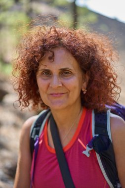 Gülümseyen orta yaşlı, kıvırcık saçlı, pembe kolsuz bir bluz giyen ve sırtında bir kamera ve sırt çantası taşıyan Chinyero ve çam ormanlarının bulanık volkanik yamacıyla arkasında Tenerife Milli Parkı 'nın