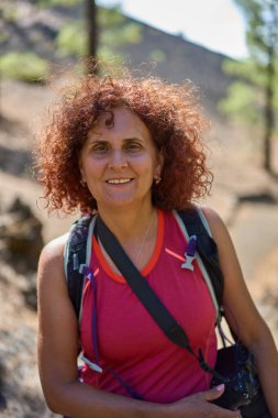 Gülümseyen orta yaşlı, kıvırcık saçlı, pembe kolsuz bir bluz giyen ve sırtında bir kamera ve sırt çantası taşıyan Chinyero ve çam ormanlarının bulanık volkanik yamacıyla arkasında Tenerife Milli Parkı 'nın