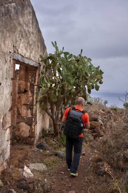 Terk edilmiş bir binanın yanından geçen sırt çantalı bir erkek yürüyüşçü ve Tenerife 'nin Anaga bölgesinde kıyı şeridi boyunca uzanan büyük dikenli armut kaktüsü. Arka planda Atlantik Okyanusu görünüyor.