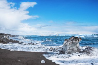 Pasifik Okyanusu 'nun güzel kış güneşli manzarası. Dalgaları yıkayan siyah volkanik kum ve buz blokları. Karla kaplı dağların manzarası, Kamçatka, Rusya.
