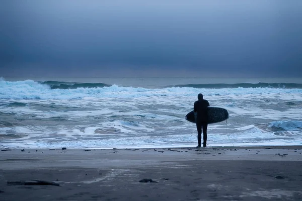 サーファーは太平洋の波が打ち寄せる前の黒い火山の砂の上に彼の手にサーフボードで立っています ロイヤリティフリーのストック写真