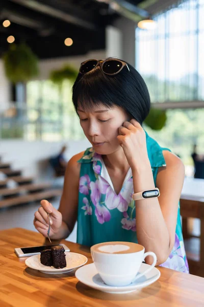 成熟したアジアの女性はコーヒーショップでチョコレートブラウニーケーキとコーヒーを食べます — ストック写真