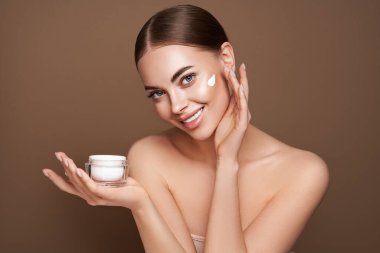 Güzellik kraliçesi yüzüne krem sürüyor. Temiz tenli genç bir kadın. Yüz kremi kavanozu olan bir model.