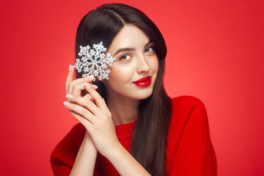 Kırmızı rujlu güzel bir genç kadın, parlak temiz bir cilt ve yüz, parlak bir kar tanesinin yüzünü gösteriyor, yüzünün yanında ve gülümsüyor, kırmızı bir arka planda soyutlanmış. Noel tatili kavramı