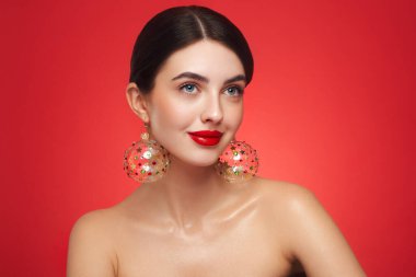 Kırmızı arka planda güzel bir genç kadının portresi, kırmızı ruj ve festival makyajı, Noel oyuncak küpeler, parlak cilt, kış tatili konsepti.