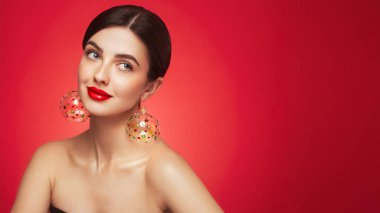 Kırmızı arka planda güzel bir genç kadının portresi, kırmızı ruj ve festival makyajı, Noel oyuncak küpeler, parlak cilt, kış tatili konsepti.