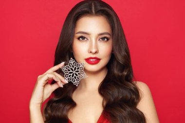 Kırmızı rujlu güzel bir genç kadın, parlak temiz bir cilt ve yüz, parlak bir kar tanesinin yüzünü gösteriyor, yüzünün yanında ve gülümsüyor, kırmızı bir arka planda soyutlanmış. Noel tatili kavramı