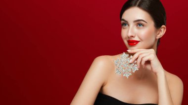 Kırmızı rujlu güzel bir genç kadın, parlak temiz bir cilt ve yüz, parlak bir kar tanesinin yüzünü gösteriyor, yüzünün yanında ve gülümsüyor, kırmızı bir arka planda soyutlanmış. Noel tatili kavramı