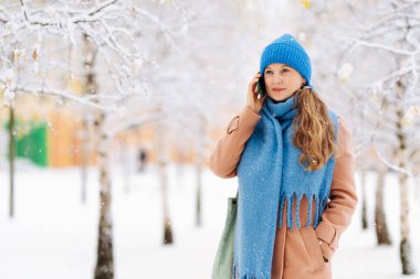 Kadın kışın bahçede yürüyor, telefonla konuşuyor. Kadın şapka ve eşarp takıyor.