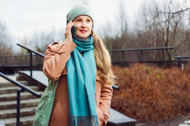 Kadın kışın bahçede yürüyor, telefonla konuşuyor. Kadın şapka ve eşarp takıyor.