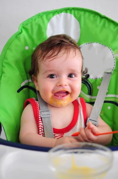 Smiling messy baby sitting in high chair holding spoon — Stock Photo, Image