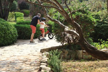 Baba, küçük çocuğuna dışarıda bisiklet sürmeyi öğretirken kucağına alıp rehberlik ediyor..