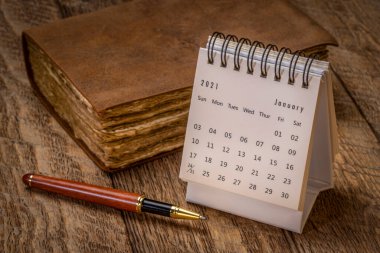 January 2021- spiral desktop calendar against rustic wood with an antique leather-bound journal and a stylish pen, season,  time and business concept clipart