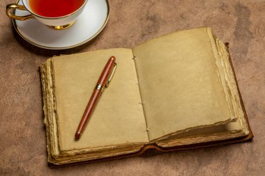 Süslü el yapımı kağıt sayfaları olan, el yapımı kağıt kaplı boş antika bir günlük. El yapımı kağıda karşı şık bir kalem ve bir fincan çay, günlük konsepti.