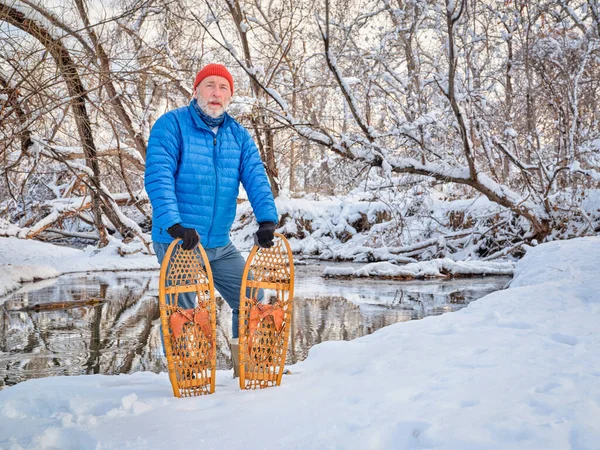 senior male with classic wooden snowshoes (Bear Paw) on a stream shore, winter scenery in Colorado