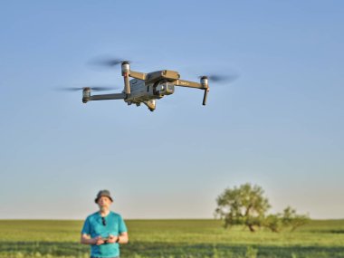 Briggsdale, CO, USA - June 8, 2021:  Radio controlled DJI Mavic 2 Pro quadcopter drone is flying over green prairie with a male pilot in background.
