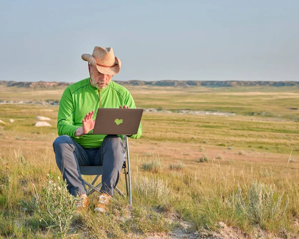 Senior man in cowboy hat is working on laptop in the middle of nowhere, early morning in Pawnee National Grassland in Colorado