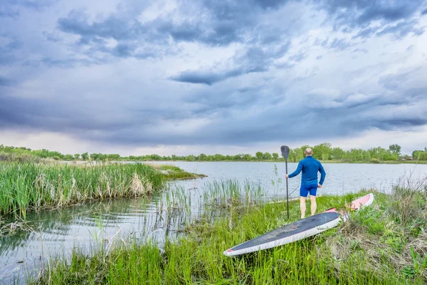 Senior paddler with SUP paddleboar watching stormy sky — Stock Photo, Image