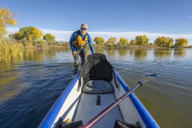 70 'li yaşlardaki yaşlı erkek raketçi, sonbahar manzarasında Colorado' da sakin, sakin bir gölde şişme bir kano fırlatıyor..