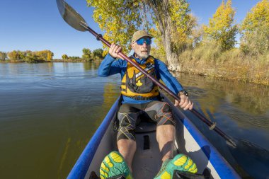 70 'li yaşlardaki yaşlı erkek raketçi, sonbahar manzarasında Colorado' da sakin bir gölde şişme bir kanoda kürek çekiyor..