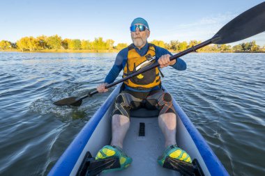 70 'li yaşlardaki yaşlı erkek raketçi, sonbahar manzarasında Colorado' da sakin bir gölde şişme bir kanoda kürek çekiyor..