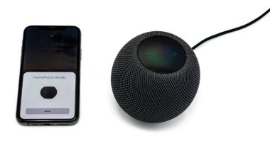 Siyah bir Apple Homepod Mini akıllı hoparlör açık beyaz arka planda gölgelerle çekilmiş Apple iPhone 12 kullanıyor..
