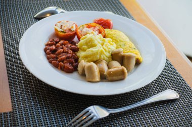 Beyaz bir tabakta servis edilen patates püresi, omlet, fasulye, domates ve sosislerden oluşan sağlıklı kahvaltı menüsü.