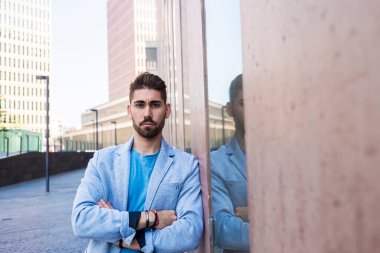 Çapraz kollu sakallı adam, modaya uygun bir model, kentsel arka planda günlük kıyafetler giyiyor.