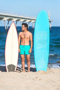 Neoprene mayo giymiş yakışıklı ve çekici bir sörfçü elinde sörf tahtası, yaz sporu ve tatillerin tadını çıkardıktan sonra havalı poz veriyor.