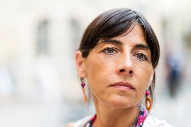 Çekici, kendine güvenen, uzun siyah saçlı, açık havada mavi gözlü, yüzünde ciddi bir ifadeyle bakan genç bir kadın.