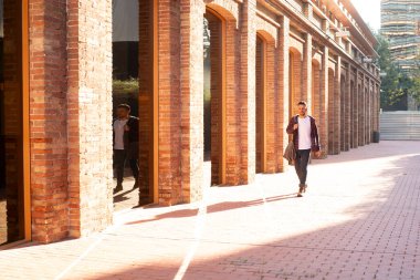 Yetişkin beyaz bir adam tuğla koridorda tek başına yürüyor. Günlük kıyafetler giyiyor, ceket giyiyor ve modern mimarinin yanında gün ışığında hareket ediyor..