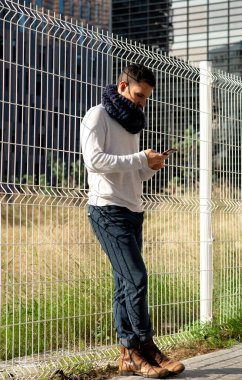 Metal bir çitin yanında duran, akıllı telefonuna bakan yetişkin beyaz bir adam. Günlük kıyafetler, kentsel ortam, gündüz manzarası..