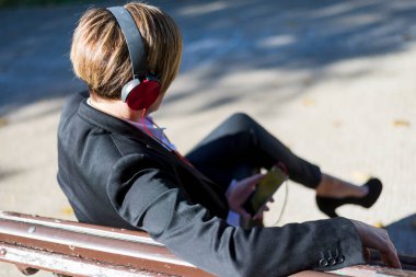 Siyah takım elbiseli yetişkin beyaz iş kadını park bankında oturuyor, kırmızı kulaklık takıyor ve açık havada akıllı telefon tutuyor..