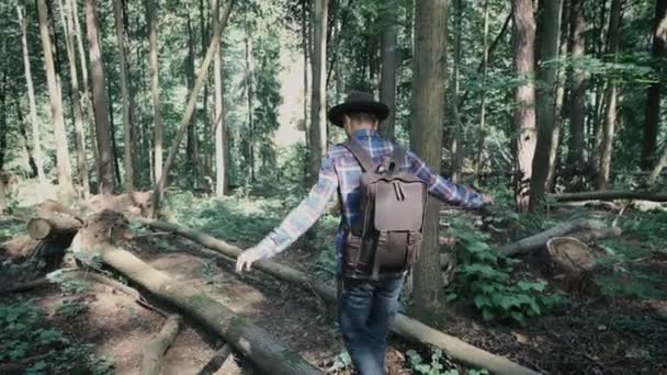 穿着毛毡帽和格子呢衬衫 背着皮背包 穿着毛毡帽的年轻男子在森林里沿着倒下的树干走着 保持着身体的平衡 冒险的激情和流浪的欲望 — 图库视频影像