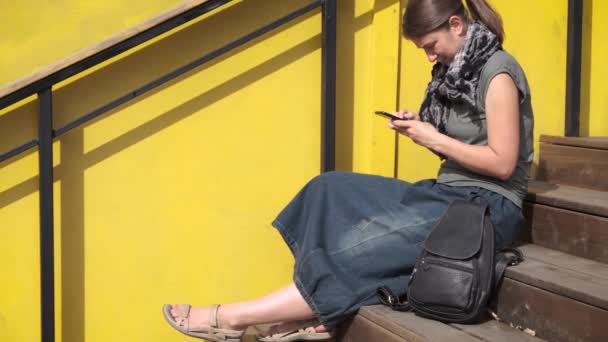 黄色の壁の近くの階段に座って スマートフォンを使用して流行の革のバックパックとブーホースタイルで身に着けている女性 ファッションアクセサリー カジュアルストリートウェア — ストック動画