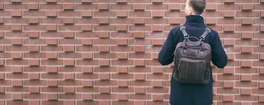 Tuğla duvarın yanında duran deri sırt çantalı, modaya uygun sakallı adam. Şık aksesuarlar, sokak giysileri, erkek şehir modası..