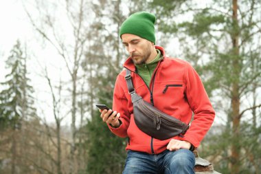 Kırmızı ceketli sakallı bin yıllık bir adamın portresi, örgü şapkalı ve deri çantalı, sonbahar parkında otururken akıllı telefon kullanıyor.. 