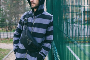 Çizgili kapüşonlu siyah deri çantalı sakallı genç bir adamın kesilmiş portresi. Sokak giysisi, moda şehri aksesuarları.. 