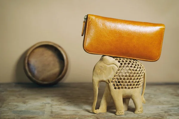 Ginger leather purse lies on back of wooden elephant. Fashion accessories in Indian folklore style. Composition in colors of nature, earth tone.