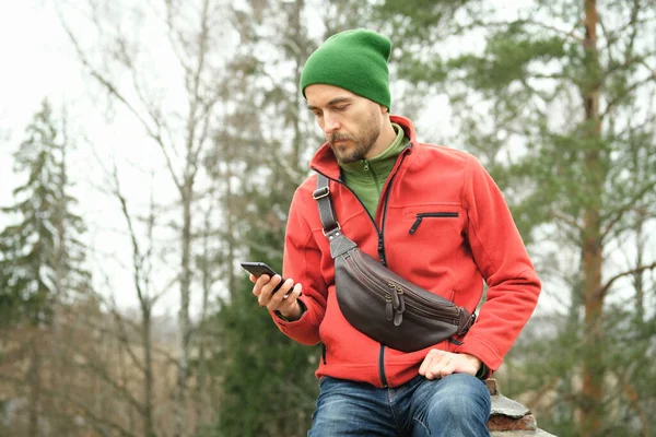 Kırmızı ceketli sakallı bin yıllık bir adamın portresi, örgü şapkalı ve deri çantalı, sonbahar parkında otururken akıllı telefon kullanıyor.. 